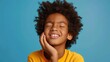 Upset handsome African American boy with closed eyes touching cheek, having toothache wearing stylish yellow casual t shirt isolated on blue background. Dental treatment concept