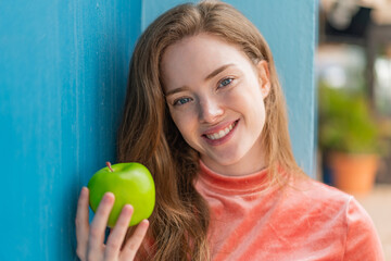 Wall Mural - Young redhead woman with an apple at outdoors smiling a lot