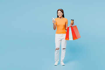 Full body fun young woman wears orange t-shirt casual clothes hold in hand paper package bags after shopping use mobile cell phone isolated on plain blue background. Black Friday sale buy day concept.