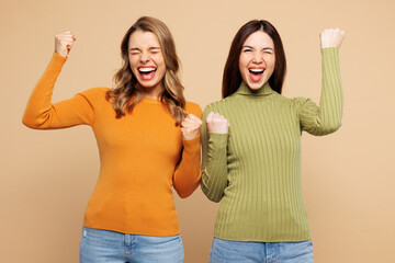 Wall Mural - Young friends two women they wear orange green shirt casual clothes together doing winner gesture celebrate clenching fists isolated on plain pastel light beige background studio. Lifestyle concept.