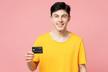 Wall Mural - Young smiling happy fun Caucasian man he wears yellow t-shirt casual clothes hold in hand mock up of credit bank card isolated on plain pastel light pink background studio portrait. Lifestyle concept.