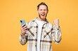 Young smiling happy Caucasian man he wear brown shirt casual clothes hold in hand use mobile cell phone chatting online do winner gesture isolated on plain yellow orange background. Lifestyle concept.