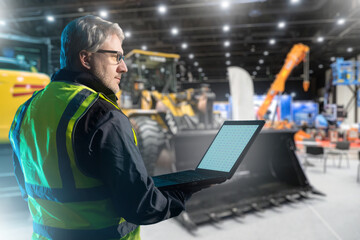 Poster - Experienced man in building with construction machines. Industrial special machines inside hangar. Gray-haired man with laptop. Worker at construction equipment exhibition. Enterprise engineer