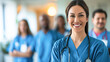 Confident female healthcare professional in scrubs with a stethoscope, team of medical staff in background.