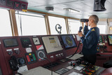 Fototapeta Do pokoju - Officer on watch with radio on the navigational bridge. Caucasian man in blue uniform sweater on the bridge of cargo ship.