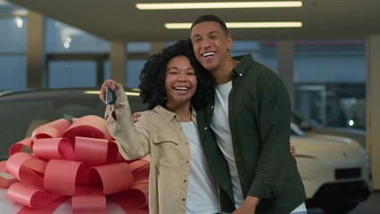 Wall Mural - Excited young happy couple in love celebrating purchase new car luxurious present with big bow on automobile hood African American woman man showing keys of auto buying in modern dealership showroom