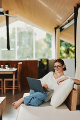 Wall Mural - Woman working on laptop in cozy home office on the couch, surrounded by modern decor She is a young freelancer, enjoying the freedom of working from home With a smile on her face and glasses perched