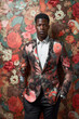 a black man wearing a floral jacket in front of a floral wallpapered wall