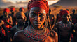 A young African woman wearing a red turban, beaded necklaces and face paint stands in front of a group of people in the background