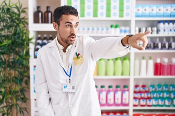 Poster - Handsome hispanic man working at pharmacy drugstore pointing with finger surprised ahead, open mouth amazed expression, something on the front