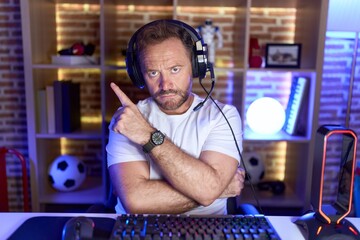 Poster - Middle age man with beard playing video games wearing headphones pointing with hand finger to the side showing advertisement, serious and calm face