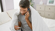 © Krakenimages.com - Handsome young hispanic man with a beard looking at a thermometer while wrapped in a blanket on his bed and talking on the phone in a bedroom.