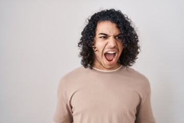 Sticker - Hispanic man with curly hair standing over white background angry and mad screaming frustrated and furious, shouting with anger. rage and aggressive concept.