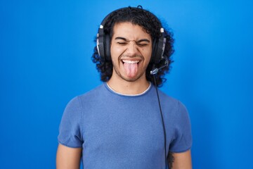 Wall Mural - Hispanic man with curly hair listening to music using headphones sticking tongue out happy with funny expression. emotion concept.