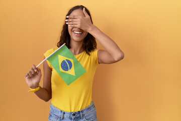 Wall Mural - Young hispanic woman holding brazil flag smiling and laughing with hand on face covering eyes for surprise. blind concept.