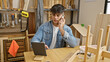 © Krakenimages.com - Hardworking young hispanic man, a dedicated builder and carpenter engaged in his profession, talking on smartphone and using touchpad amidst the lumber and timber at his workshop