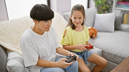 Poster - Happy mother and daughter sitting on a comfortable sofa, playing video game, smiling together at home