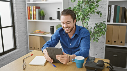 Wall Mural - Young hispanic man business worker using touchpad smiling at the office