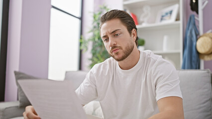 Wall Mural - Young hispanic man reading document sitting on sofa at home