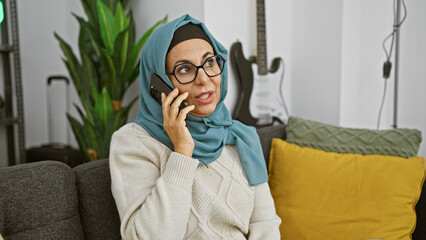 Sticker - A mature woman in a hijab talking on the phone while seated on a sofa in a modern living room.