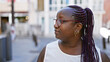 Cool plus size african american woman in glasses, with braids, casting a serious expression while looking to the side on a sunny urban street.