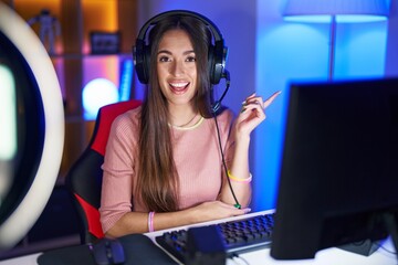 Canvas Print - Young hispanic woman playing video games with a big smile on face, pointing with hand and finger to the side looking at the camera.