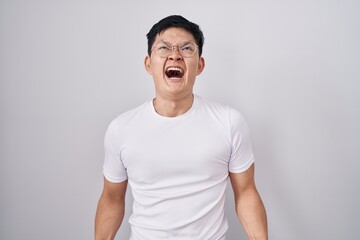 Sticker - Young asian man standing over white background angry and mad screaming frustrated and furious, shouting with anger. rage and aggressive concept.