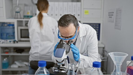 Poster - Two dedicated scientists, team mates working together indoors in a lab, busy with serious research using a microscope, discovering new analysis in medical science.