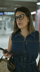 Poster - Stunning hispanic woman, glasses on, standing at underground railroad station, anticipating her subway journey, engrossed in her phone