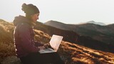 Fototapeta  - Woman working on laptop in sunset mountains. Young freelancer tourist caucasian female typing on notebook sitting on autumn grass meadow landscape. Concept of remote online work, study or business
