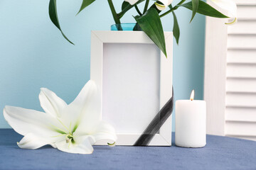 Wall Mural - Blank funeral frame, burning candle and vase with lily flowers on table near color wall, closeup