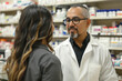 © nudjaree - Mature male pharmacist working with female customer in drugstore. Mature man in white coat and eyeglasses standing in drugstore. Pharmacy concept