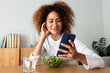 © Daniel - Young multiracial woman eating healthy salad while using phone at home