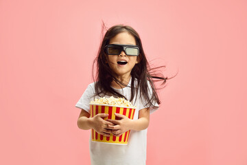 Joyful and Surprised Child Girl in 3D Glasses Holding Popcorn Bucket Having Flowing Hair Standing Over Pink Isolated Background.