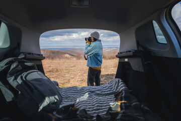 Wall Mural - Young happy woman sitting in an open car trunk taking pictures with a camera. Travel by car