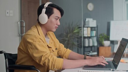 Side view of Asian wheelchaired coder listening to music in headphones while developing app on laptop in office