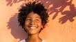 A young child with curly hair smiling brightly standing in front of a textured orange wall with shadows of leaves.