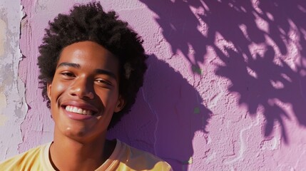 Wall Mural - A young man with a radiant smile and curly hair standing against a vibrant purple wall bathed in soft sunlight.