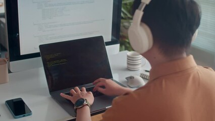 Back view of male programmer in headphones coding on laptop sitting in front of big monitor at office