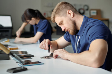 Poster - Young male technician or repairman bending over desk and fixing tiny details of smartphone with screwdriver against female colleague