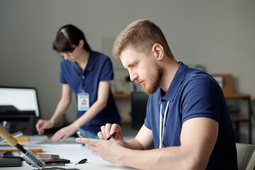 Poster - Young technician checking battery of broken mobile phone while sitting by workplace in repair center office and identifying technical problem