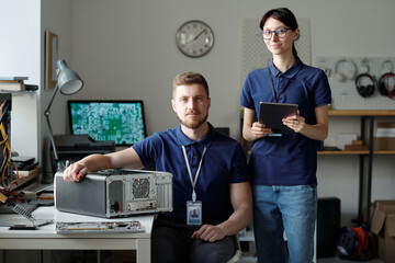 Poster - Team of two young successful engineers of repair service center in blue uniform looking at camera while one of them using tablet
