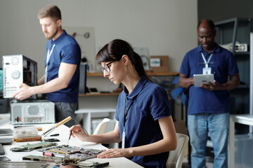 Poster - Young female technical service specialist fixing details of disassembled laptop with screwdriver against intercultural male colleagues