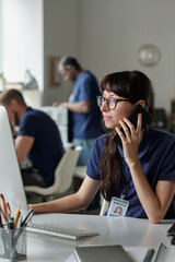 Poster - Young businesswoman in eyeglasses looking at computer screen and clicking mouse while talking to client on mobile phone by workplace