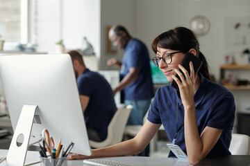 Poster - Young smiling female technician in eyeglasses communicating to client on cellphone and looking at computer screen in coworking space