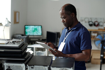 Poster - Serious mature African American technician with tablet standing in front of camera by workplace and looking through online manual