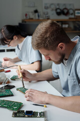 Poster - Young man with electric handtool bending over desk and motherboard while repairing detail of computer processor against colleague