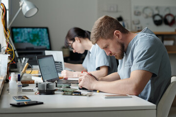 Poster - Professional young technician bending over desk while repairing or checking detail of computer motherboard against female colleague