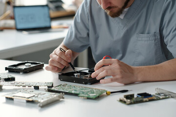 Poster - Cropped shot of young man repairing detail of computer processor with electric transmission tweezers while sitting by workplace