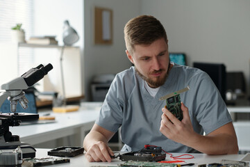 Poster - Young serious man in grey uniform looking at part of computer processor while sitting by workplace and identifying technical problem